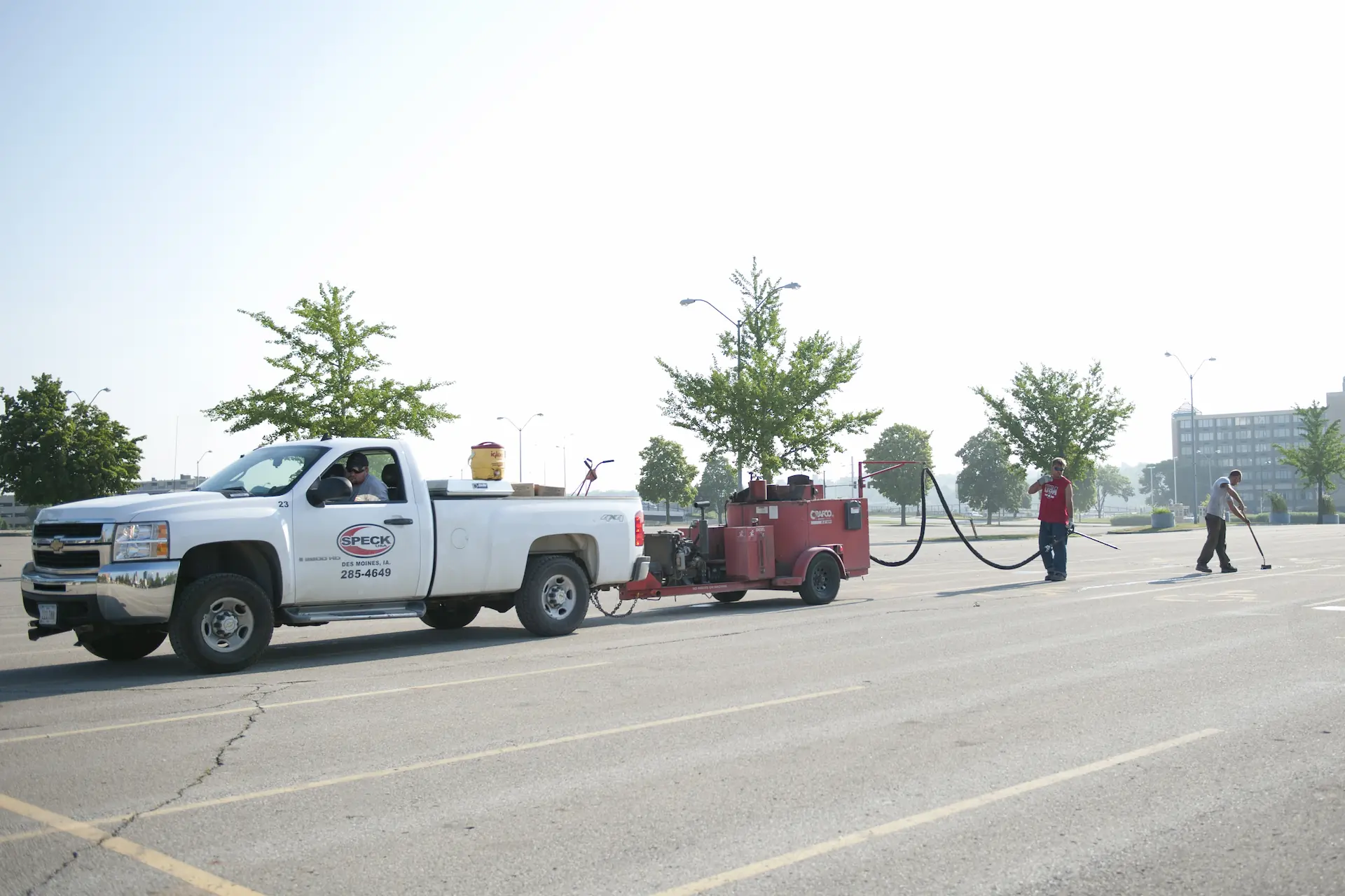 A white pickup truck from Speck USA with a resurfacing vehicle in tow repairs a commercial parking lot. Resurfacing and sealing from professional-grade services like these are part of how to maintain your parking lot.