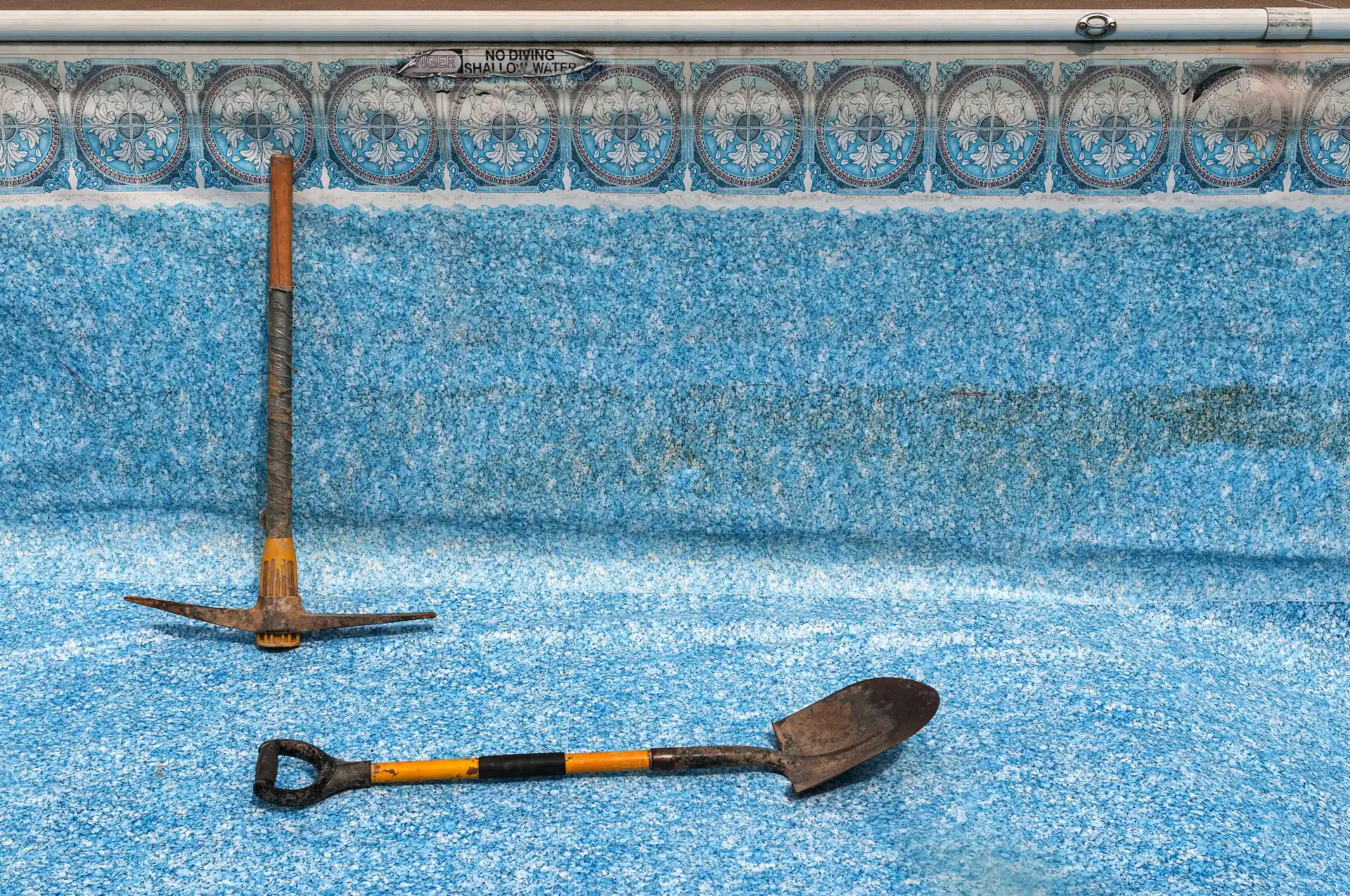 Medium shot of a pick axe and shovel sitting in an empty inground pool with a faded, discolored vinyl pool liner. Discoloration and stains are signs that it's time to replace your vinyl liner.