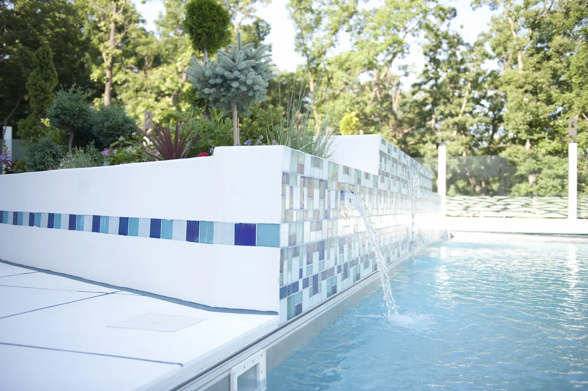 Side shot of a concrete pool with a contemporary white deck and cascades pouring water from the pool wall. Cascades are one feature that can be incorporated into a concrete pool design.