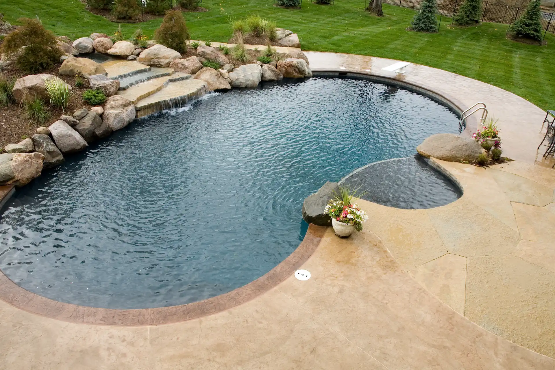 Wide, aerial shot of a kidney-shaped inground pool with a jade-colored liner decorated with river rocks. Naturalistic colors and features like the kind pictured here are among the popular pool design trends for 2023.