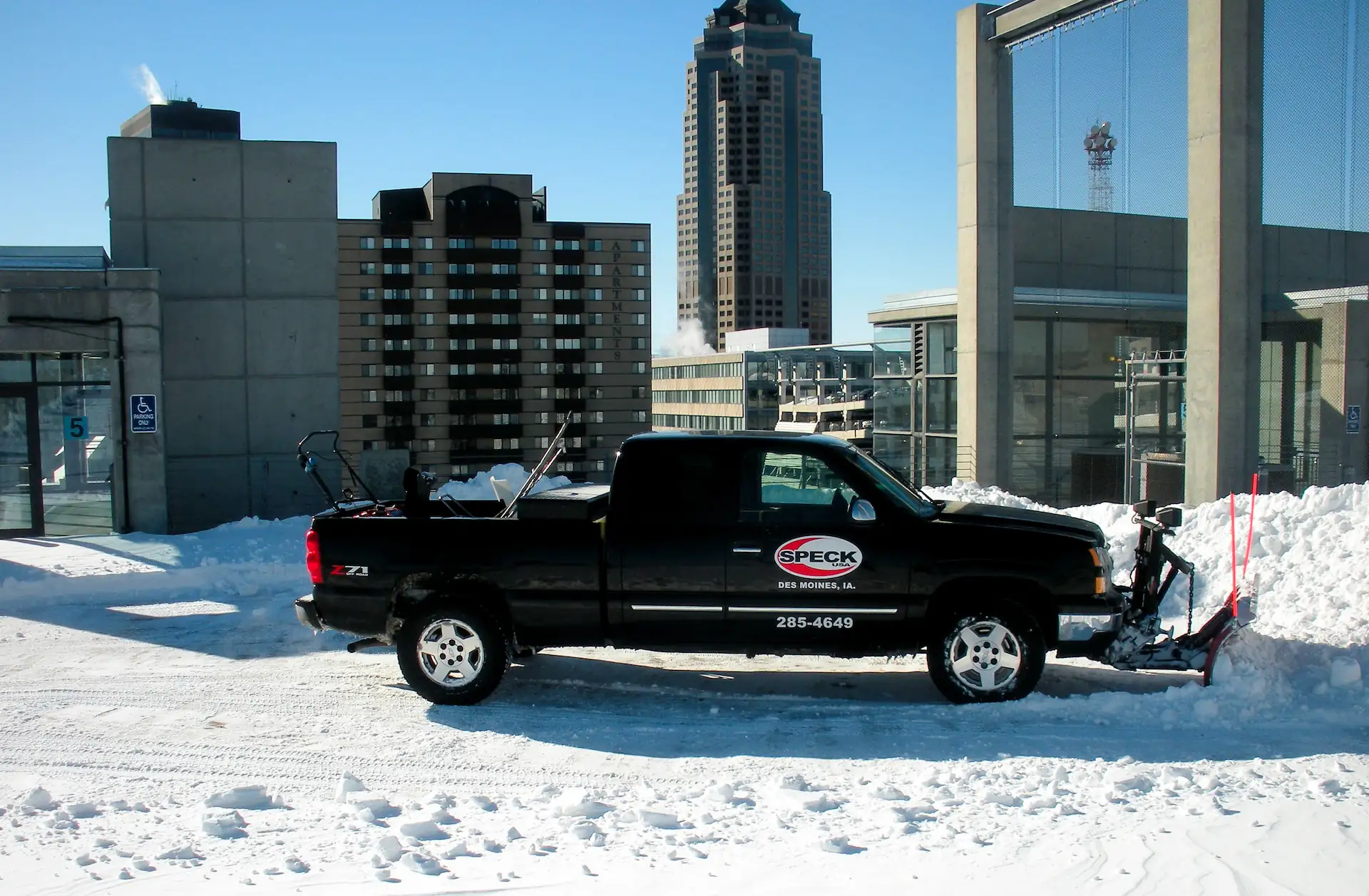 A black pick up truck with the Speck USA logo on the side and a snow plow attached to the front sits in a snow-covered parking lot in the daylight. Hiring snow removal services is an important part of maintaining business in winter.